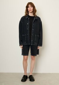 Overmaat zwarte denim jacket met twee voorzakken, gecombineerd met zwarte denim shorts. Model draagt zwarte instappers. Effen achtergrond.