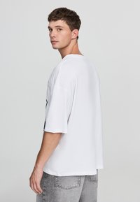 Biała koszulka z krótkim rękawem i okrągłym dekoltem. Wykonana z miękkiej bawełny, luźny krój, gładki wzór. Szare dżinsy i neutralne tło.