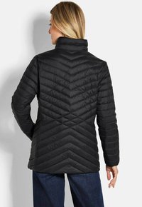 Vrouw met blond haar die een zwarte gewatteerde jas met chevronstiksels draagt, staande met haar rug naar de camera tegen een witte achtergrond.