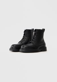Un par de botas de tobillo de cuero negro con cordones, cremallera lateral y suelas de goma gruesa sobre un fondo claro y sencillo.