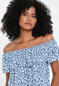 Ung kvinde med krøllet hår iført en blå off-shoulder kjole med hvide blomster mønstre, som ser selvsikkert ind i kameraet.
