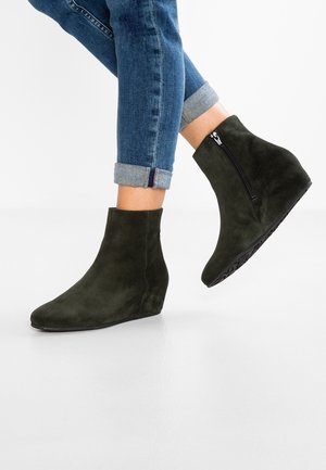 Bottes à talon compensé en suede vert avec un bout rond et une fermeture éclair sur le côté, sur un fond blanc lisse. Les jeans sont retroussés aux chevilles.