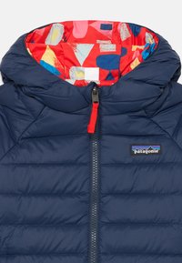 Marineblaue Pufferjacke mit hohem Kragen, Reißverschluss vorn und roten Akzenten. Das Innere zeigt ein farbenfrohes geometrisches Muster. Patagonia-Logo auf der Brust.