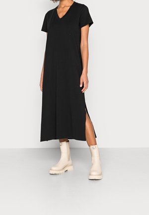 Frau trägt ein schwarzes, kurzärmeliges V-Ausschnitt-Midikleid mit Schlitz an der Seite und beige klobige Stiefeletten, steht vor einem einfarbigen Hintergrund.