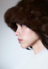 Giovane donna di profilo con un cappello di pelliccia marrone scuro e un indumento bianco e soffice, contro uno sfondo semplice.