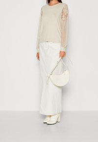 Beżowa bluzka z długim rękawem z koronkowymi rękawami, zestawiona z białą długą spódnicą dżinsową i białymi botkami na słupku. Trzyma białą torbę.