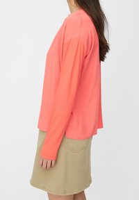 Korálová halenka s dlouhým rukávem a volným střihem, vyrobená z hladkého materiálu; kombinována s béžovou sukní ke kolenům, která má zadní kapsu.