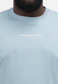 Jasnoniebieska koszulka z krótkim rękawem z bawełny, z napisem "Calvin Klein Jeans" oraz "ZAŁOŻONA W TYSIĄC SIEDMIUSET OSIEMDZIESIĄTYM" poniżej, w białym kolorze.