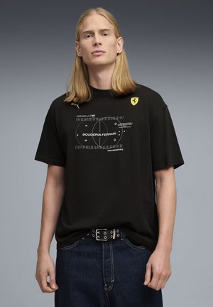 Svart bomulls-T-shirt med korta ärmar, vit grafisk tryck och Scuderia Ferrari-logotyp på övre vänstra sidan. Bärs med mörkblå jeans.