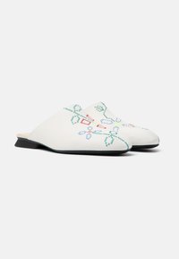 Vita slip-on-skor med en låg svart klack, prydda med grön och röd blomsterbroderi och kontrasterande sömnad längs kanterna.