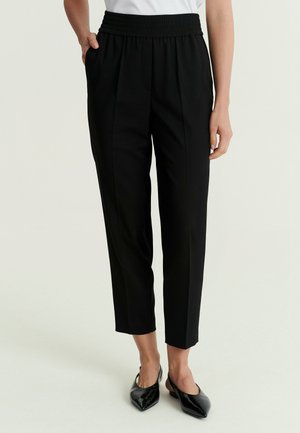 Femme portant un pantalon noir sur mesure coupé court avec une taille élastique et des chaussures slingback noires à bout pointu.