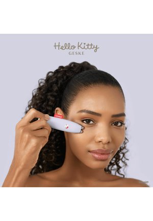 Frau verwendet einen Hello Kitty Gesichtsroller auf ihrer Wange vor einem einfarbigen hellvioletten Hintergrund.