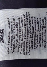 Bijela tkaninska oznaka s crnim tekstom koja navodi informacije o proizvodnji na više jezika. Sadrži QR kod u gornjem lijevom kutu.