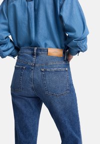 Jean en denim bleu à taille mi-haute, avec deux poches arrière et une étiquette en cuir sur la ceinture. Fini au tissu texturé.