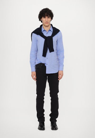 Helles blaues Hemd mit einem subtilen Muster, kombiniert mit schwarzen Skinny-Jeans und schwarzen Schuhen, dazu ein marineblauer Pullover lässig über die Schultern drapiert.