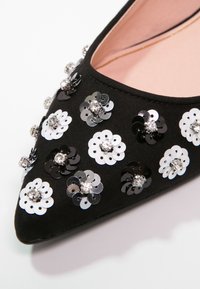 Chaussure plate pointue noire décorée d'appliques fleurs à sequins noirs et blancs alternés, chacune avec un strass central.