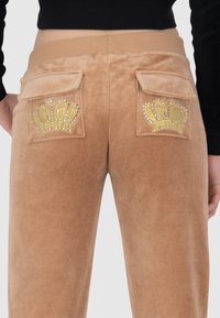 Tan velours broek met twee achterzakken voorzien van gouden kroon ontwerpen versierd met rhinestones. Zachte textuur, taps toelopende pasvorm.