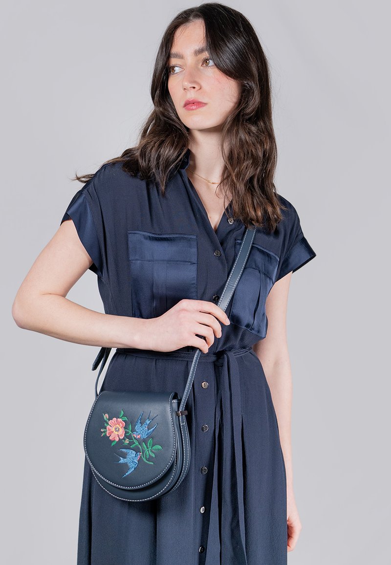 Dunkelblaue Leder-Umhängetasche mit floralen und vogelartigen Stickereien, ovale Form, glatte Textur und verstellbarer Tragegurt.