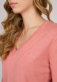 Pull en V rose en tissu tricoté doux. Doté d'un logo brodé discret sur le côté gauche. Manches longues, légèrement amples.