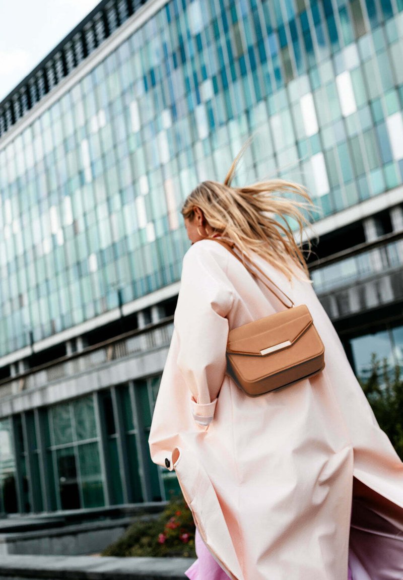 Lichtroze oversized jas met opgerolde mouwen, gecombineerd met een tan crossbody tas met een metalen accent. Modern stedelijk achtergrond.