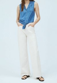 Jasnoniebieska denimowa koszula bez rękawów związaną z przodu, zestawiona z kremowymi spodniami o szerokich nogawkach i czarnymi klapkami, prezentowana na jasnym tle.