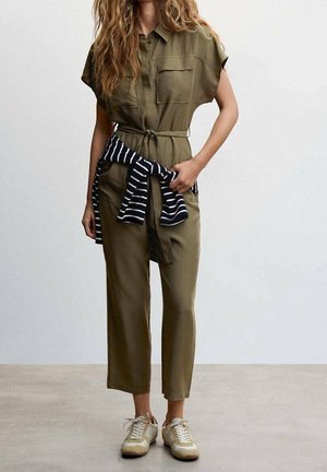 Combinaison vert olive à manches courtes, taille cintrée, poches avant, associée à un pull rayé noir et blanc noué à la taille. Baskets blanches.