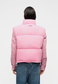 Άτομο με ίσια σκούρα μαλλιά που φοράει ένα ροζ μαξιλαρωμένο γιλέκο με το κείμενο "American Vintage", ροζ μανίκια μακριά και μπλε τζιν, γυρισμένο με την πλάτη.