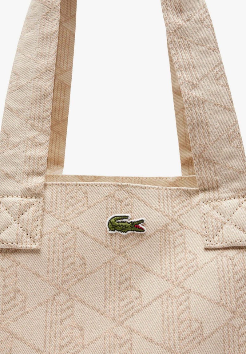 Beige Tragetasche mit geometrischem Muster, mit eingenähten Griffen und einem kleinen grünen Krokodil-Logo in der Mitte nahe oben.