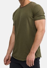 Camiseta de manga corta verde oliva con escote redondo, que presenta un corte relajado y una textura suave, con un dobladillo trasero ligeramente más largo.