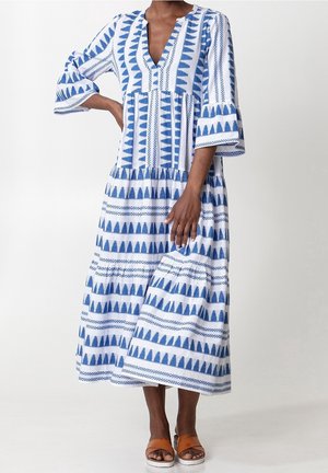 Femme portant une longue robe blanche et bleue à motifs avec manches trois-quarts et des sandales marron à bouts ouverts, debout avec une main sur la hanche.