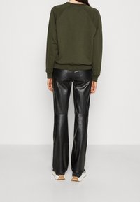 Donna che indossa un felpa verde scuro, pantaloni di pelle neri e sneakers bianche, in piedi contro uno sfondo chiaro e semplice, vista posteriore.