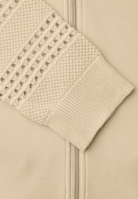 Rękaw kremowego, dzianinowego swetra z teksturowanymi wzorami spoczywający na beżowym tle z tkaniny z widocznym zamkiem błyskawicznym.