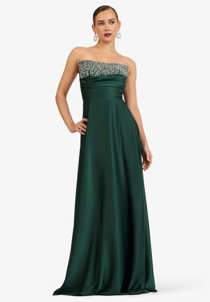 Woman in a strapless dark green satin gown with beaded silver embellishments on the bodice, posing against a white background.