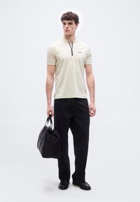 Νεαρός άνδρας με μπεζ κοντομάνικο πουκάμισο με φερμουάρ, μαύρο παντελόνι, μαύρα παπούτσια, κρατώντας μια μεγάλη μαύρη τσάντα στο δεξί του χέρι, όρθιος.