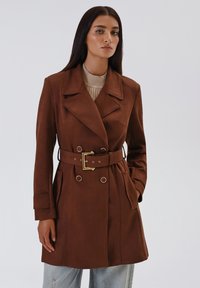 Cappotto trench marrone con design doppiopetto, dotato di colletto a risvolto, due tasche frontali e dettagli in metallo dorato.