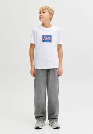 Blond jongetje staand, draagt een wit Jack & Jones T-shirt, losse grijze spijkerbroek en grijze sneakers, kijkt naar links tegen een effen achtergrond.