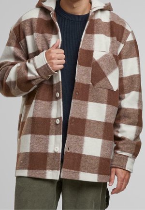 Mann trägt eine braun-weiß karierte Jacke über einem dunkelblauen Pullover mit grüner Hose und richtet den Jackenkragen.