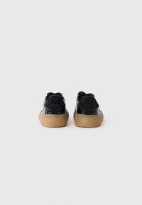 Une paire de baskets noires avec des détails en daim et en cuir verni, semelles texturées en caoutchouc gomme, montrées de l'arrière sur un fond blanc.