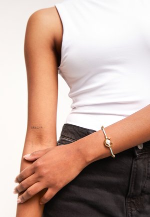 Pulsera de oro y turquesa con un adorno circular y detalle de perla, llevada en una mano que descansa sobre una prenda de mezclilla negra. El tono de piel es medio.