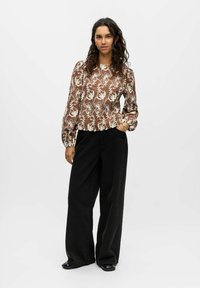 Bruine bloemen blouse met lange mouwen en een crèmekleurig patroon, gecombineerd met wijde zwarte broek. De blouse heeft een ronde halslijn en gatherende mouwen.