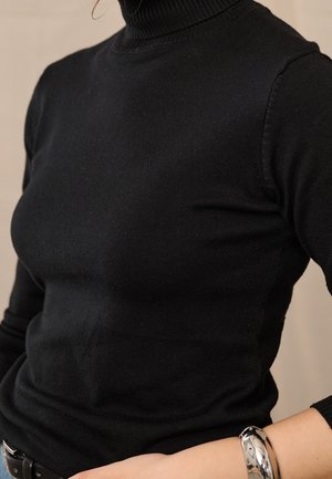 Persoon draagt een getailleerde zwarte coltrui met een zilveren armband om de pols tegen een neutrale achtergrond.