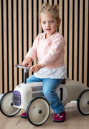 Mädchen mit blonde Haare, trägt einen rosa Pullover und Jeans, sitzt auf einem weißen Spielzeug-Pedalauto mit der Nummer 1, drinnen vor einer Holzpaneelwand.