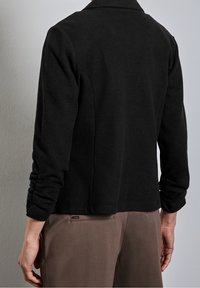 Svart jacka med långa ärmar, mjuk textur och krage. Har en figursydd design och lätt skräddarsydda ärmar.