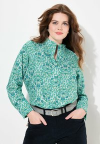 Camisa verde de botones con patrón floral, incluyendo tonos de azul y verde, y un cuello tipo banda. Combinada con un cinturón gris y pantalones oscuros.