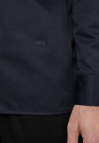 Marinblå jacka i slät tyg, med en diskret broderad detalj "W.W." nära fållen. Strukturerad med en ren finish.