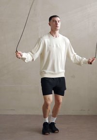 Hvid sweatshirt med præget logo, sorte shorts, hvide sokker og sorte sneakers. Mand, der hopper i sjippetov mod en ensfarvet baggrund.