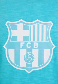Tissu bleu clair arborant un blason blanc du FC Barcelone avec un motif rayé, une croix et un ballon de football au centre.