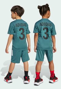 Completo da allenamento turchese composto da maglia e pantaloni corti, caratterizzato dal logo "ADIDAS" nero e dal numero 3 con un design a ragnatela. Sneakers nere e rosse.
