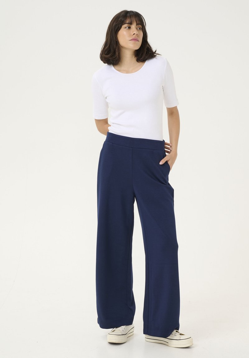 Marineblauwe wijde pijpenbroek met een gladde textuur en zijzakken, gecombineerd met een aansluitend wit T-shirt met korte mouwen. Model staat op een effen achtergrond.