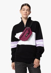 Borsa a vita bordeaux con chiusura con zip, indossata in diagonale sopra una felpa a righe bianche e nere. La borsa presenta un patch con il logo.